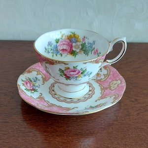 Könnte beinhalten: Ein Vintage-Tee-Set, bestehend aus einer Tasse und einer Untertasse. Die weiße Porzellantasse und Untertasse haben eine rosa-goldene Verzierung mit floralen Mustern. Die Tasse hat einen Henkel und steht auf einem Sockel.