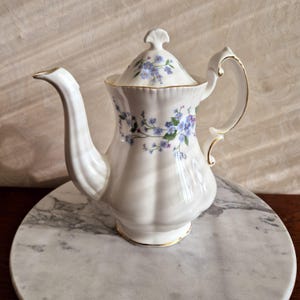 May include: A white porcelain teapot with a floral design featuring blue forget-me-not flowers. The teapot has a gold trim around the lid and base.