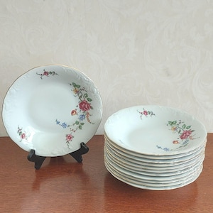 Könnte beinhalten: Ein Set aus weißen Porzellansuppentellern mit einem zarten Blumenmuster aus roten, gelben und blauen Blüten sowie grünen Blättern. Jeder Teller hat einen gewellten Rand mit einem dünnen Goldrand und geprägten Details am äußeren Rand.