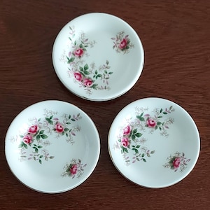 May include: Three white ceramic saucers with a gold rim and a floral design featuring pink roses and green leaves.