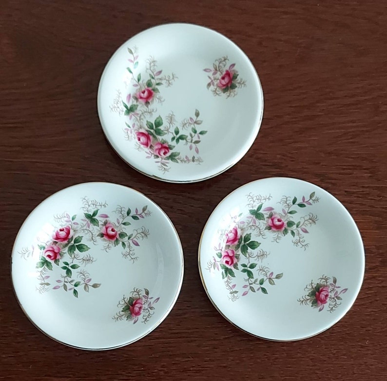 May include: Three white ceramic saucers with a gold rim and a floral design featuring pink roses and green leaves.
