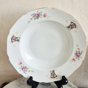 May include: White porcelain plate with a scalloped edge and gold trim. The plate features floral designs in pink, blue, and purple. Decorative black and gold accents are also present. The plate is displayed on a black stand.