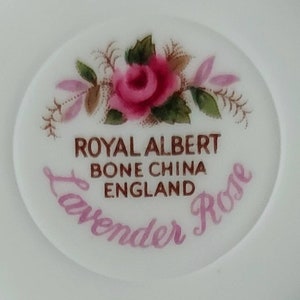 May include: A close-up of a white porcelain plate with a pink rose and green leaves. The plate has the text "Royal Albert Bone China England Lavender Rose" printed on it.