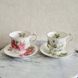 Può includere: Due tazze da tè e piattini in porcellana. Una tazza presenta disegni floreali rosa, l'altra disegni floreali bianchi. Entrambe hanno finiture e manici dorati. I piattini corrispondono ai disegni delle tazze.