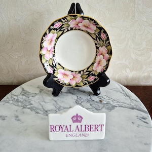 Puede incluir: Un plato decorativo con un diseño floral, con flores rosas sobre un fondo negro y un borde dorado. El plato se exhibe sobre un soporte negro. Un letrero blanco con el texto "ROYAL ALBERT ENGLAND" está frente al plato.