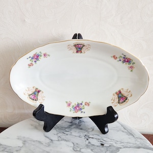 May include: An oval, cream-coloured serving platter with a gold rim and floral accents. The platter features clusters of pink, purple, and blue flowers, and decorative black and gold designs. It is displayed on a black stand.