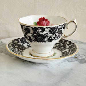Puede incluir: Juego de taza y platillo blancos con detalles de encaje negro y ribete dorado. La taza de té presenta una ilustración de rosa roja en el interior. El platillo refleja el diseño de encaje. Un juego de té clásico y elegante.