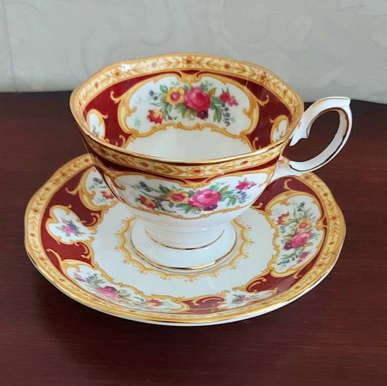 May include: A white teacup and saucer with a red and gold floral pattern. The cup has a scalloped rim and a handle. The saucer has a matching pattern.