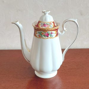 Może przedstawiać: Biały dzbanek do kawy z porcelany o karbowanym korpusie i pasującej pokrywce. Górna część ozdobiona jest ciemnoczerwonym pasem, złotymi wykończeniami oraz kolorowym wzorem kwiatowym w odcieniach różu, żółci i zieleni.