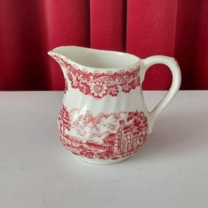 Może przedstawiać: Biały dzbanek ceramiczny z czerwono-białym wzorem kwiatowym. Dzbanek ma uchwyt i dzióbek.