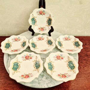 Puede incluir: Juego de cinco platos de postre de porcelana blanca con borde festoneado. Cada plato presenta un diseño floral con detalles dorados y un borde azul verdoso.