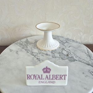 May include: A white porcelain cake stand with gold trim. The stand features a fluted base and a scalloped edge. A Royal Albert England sign is in the foreground.