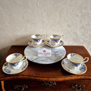 Op de afbeelding: Vier Royal Albert kopjes en schotels met een blauw-paars bloemmotief en gouden rand. De kopjes en schotels staan op een houten oppervlak en een marmeren dienblad. Een bordje toont "Royal Albert England".