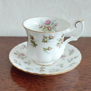 May include: A white teacup and saucer set with a floral pattern of pink and blue roses. The set has a gold trim around the edges.