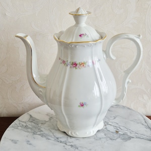 May include: White porcelain coffee pot with a lid, handle, and spout. The pot is decorated with gold trim and floral patterns in pink, blue, and yellow. The pot is sitting on a marble surface.