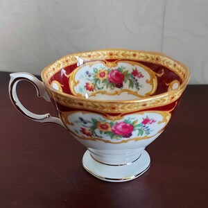 May include: A white teacup with a gold and red floral pattern. The cup has a handle and a pedestal base.