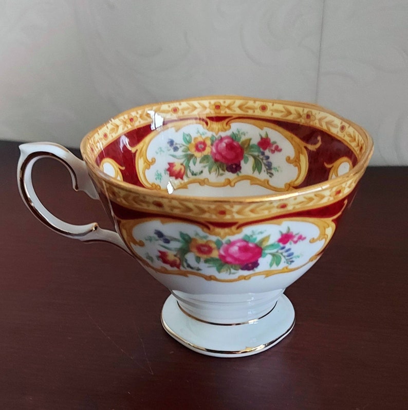 May include: A white teacup with a gold and red floral pattern. The cup has a handle and a pedestal base.