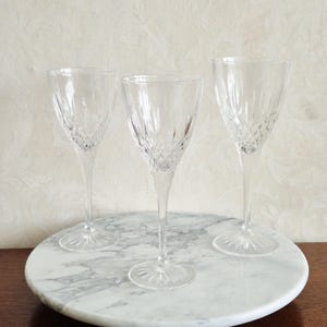 May include: Three clear crystal wine glasses with diamond-cut patterns, displayed on a round marble serving tray. The glasses have long stems and starburst bases. The background is a light-coloured wall with a subtle pattern.