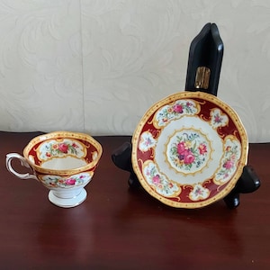 May include: A white teacup and saucer with a red and gold floral pattern. The saucer is on a black stand.