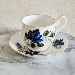 Peut inclure: Service à thé blanc avec un motif de roses bleues et des bordures dorées. La tasse a une anse incurvée et repose sur une soucoupe assortie. L'ensemble est posé sur une surface en marbre.