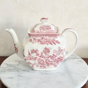 May include: A cream-colored ceramic teapot featuring a square-shaped body and a matching lid. The teapot is decorated with a detailed pink floral pattern that includes blooming flowers, foliage, and birds, with a curved handle and spout.