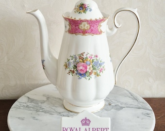 Pode incluir: Um bule de porcelana branca com um design de buquê de flores, uma faixa rosa estampada perto do topo e bordas douradas. A tampa tem um motivo floral correspondente. Uma placa retangular exibe uma coroa e o texto ROYAL ALBERT ENGLAND.