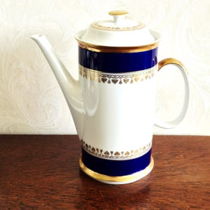 Puede incluir: Una cafetera de porcelana blanca con un pico largo, asa y tapa. La cafetera presenta una banda azul marino con detalles de pan de oro y ribete dorado. La cafetera está sobre una superficie de madera.