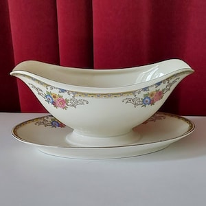 以下が含まれることがあります： ピンク、ブルー、イエローの色の花柄が施された白い磁器製のグレービーボート。ボートは金色の縁取りが施され、同じ模様のソーサーの上に置かれています。