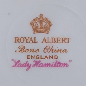 May include: A close-up of a Royal Albert Bone China teacup with a gold crown and the words "Lady Hamilton" in pink.