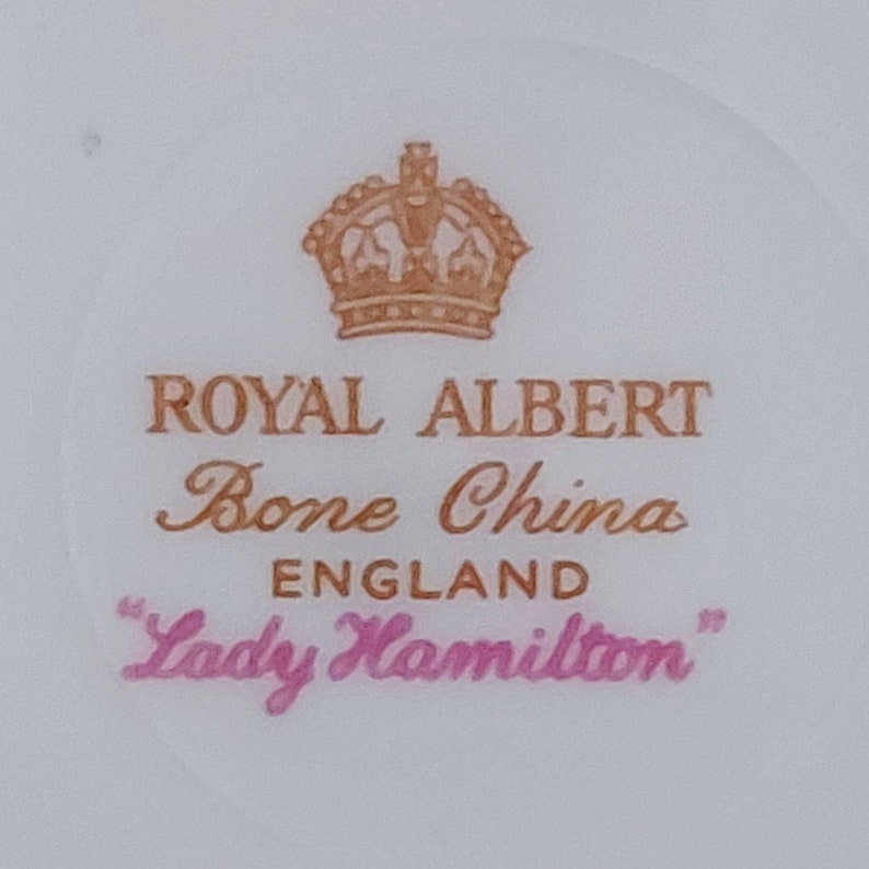 May include: A close-up of a Royal Albert Bone China teacup with a gold crown and the words "Lady Hamilton" in pink.