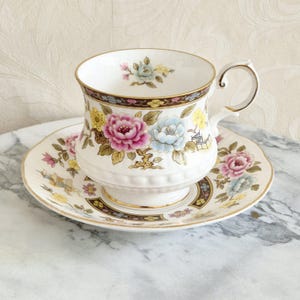 Pode incluir: Conjunto de chá branco com uma chávena e pires com um design floral com flores rosa, azuis e amarelas. A chávena e o pires têm acabamentos dourados e uma borda decorativa. O conjunto está sobre uma superfície de mármore.