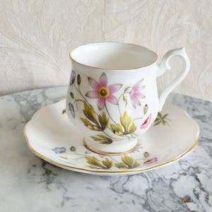 May include: White teacup and saucer set with a floral design featuring pink, yellow, and blue flowers. The cup and saucer have gold trim. The teacup has a curved handle.