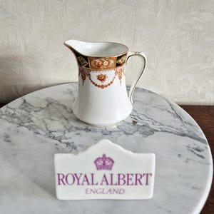 May include: A white porcelain creamer with a decorative floral and gold trim design. The creamer has a curved handle and a spout. A Royal Albert England sign is in the foreground.