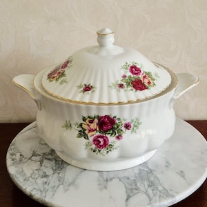 May include: A white porcelain soup tureen with a lid, adorned with a floral rose pattern in shades of red, pink, and yellow. The tureen has gold trim and handles, and sits on a marble-patterned plate.