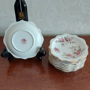 May include: A set of 10 white saucers with a gold rim and a floral pattern featuring pink roses and green leaves. The saucers are made by Royal Albert and are in the "Old Country Roses" pattern.