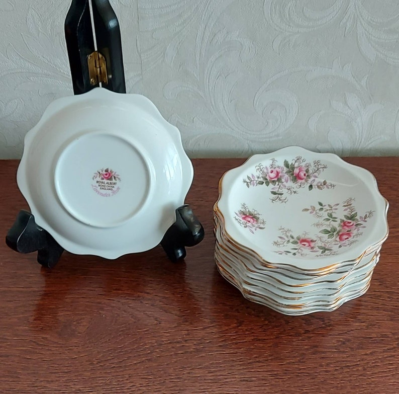 May include: A set of 10 white saucers with a gold rim and a floral pattern featuring pink roses and green leaves. The saucers are made by Royal Albert and are in the "Old Country Roses" pattern.