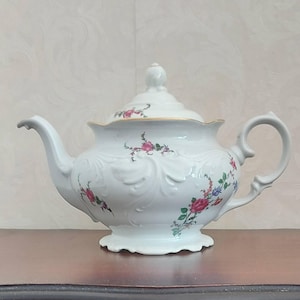 Könnte beinhalten: Eine weiße Porzellanteekanne mit einem zarten Blumenmuster aus rosa Rosen und kleinen blauen Blüten. Die Teekanne verfügt über ein geprägtes Schnörkeldesign auf dem Körper, einen gewellten Boden, einen geschwungenen Henkel und einen Goldrand.