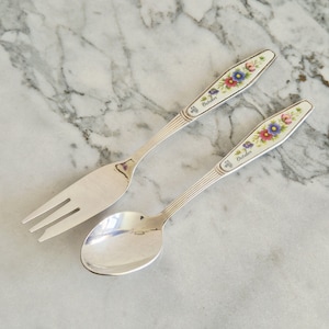 May include: A silver fork and spoon set. The handles are white with a floral design in pink, blue, and yellow. The word "October" is printed on the handle. The set is on a marble surface.