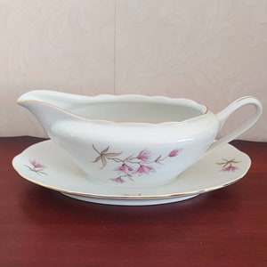Può includere: Salsiera in porcellana bianca con un motivo floreale rosa e un sottopiatto abbinato. La salsiera ha un bordo dorato e il sottopiatto ha una fascia dorata attorno al bordo.