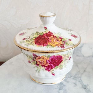 May include: A white ceramic sugar bowl with a lid, adorned with red and yellow roses and green leaves. The bowl and lid are trimmed with gold. The lid has a small, rounded handle.