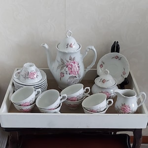 Könnte beinhalten: Ein weißes Teeservice mit rosafarbenen Blumendesigns. Das Set beinhaltet eine Teekanne, eine Zuckerdose, eine Milchkännchen und sechs Teetassen und Untertassen.