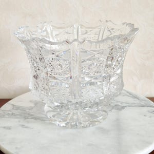 Puede incluir: Un jarrón de cristal tallado transparente con un borde festoneado y una base de pedestal. El jarrón presenta intrincados patrones geométricos, incluyendo estrellas y formas de diamante, creando un efecto brillante. El jarrón se utiliza probablemente con fines decorativos.