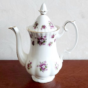 Puede incluir: Tetera de porcelana blanca con un diseño floral con flores moradas. La tetera tiene una tapa con un borde dorado y un asa.