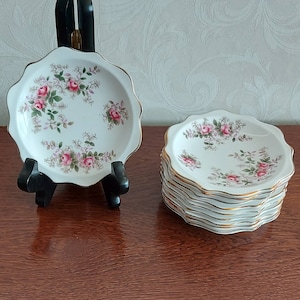 May include: A set of eight white ceramic saucers with a gold rim and a floral pattern featuring pink roses and green leaves.