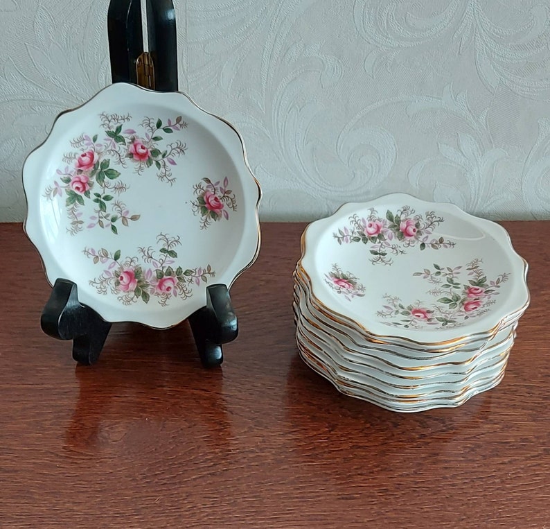 May include: A set of eight white ceramic saucers with a gold rim and a floral pattern featuring pink roses and green leaves.