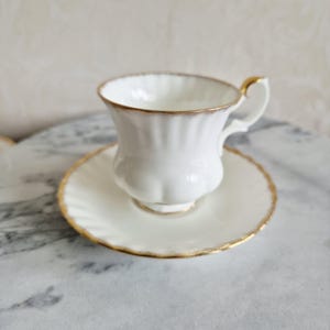 May include: A white teacup and saucer set with gold trim. The teacup has a fluted design and a curved handle. The saucer is round and also features gold trim. The set is placed on a marble surface.