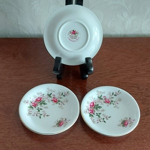 May include: Two white porcelain saucers with a floral pattern of pink roses and green leaves. The back of one saucer shows the Royal Albert logo and the words "Lavender Rose" in gold lettering.