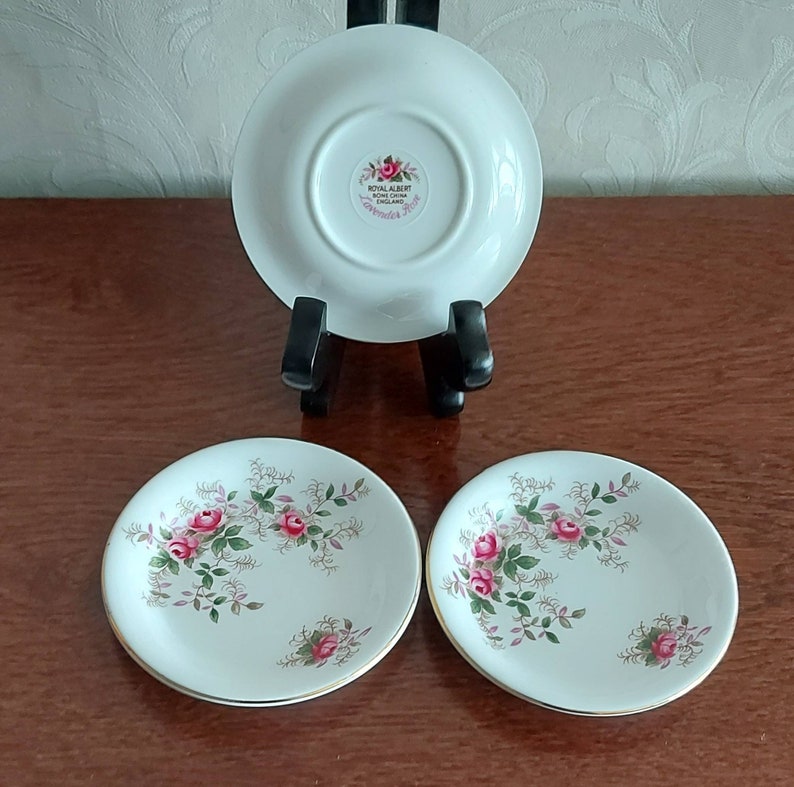 May include: Two white porcelain saucers with a floral pattern of pink roses and green leaves. The back of one saucer shows the Royal Albert logo and the words "Lavender Rose" in gold lettering.