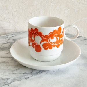 Può includere: Set tazza e piattino da caffè in ceramica bianca. La tazza presenta un vivace motivo floreale arancione. Il piattino è rotondo e bianco. La tazza ha un manico ed è appoggiata sul piattino. Il set è su una superficie di marmo.