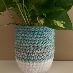 May include: A crocheted plant pot in shades of blue, gray, and white. The pot is cylindrical with a rounded base and holds a green plant with variegated leaves. The crochet pattern is visible, adding texture to the design.